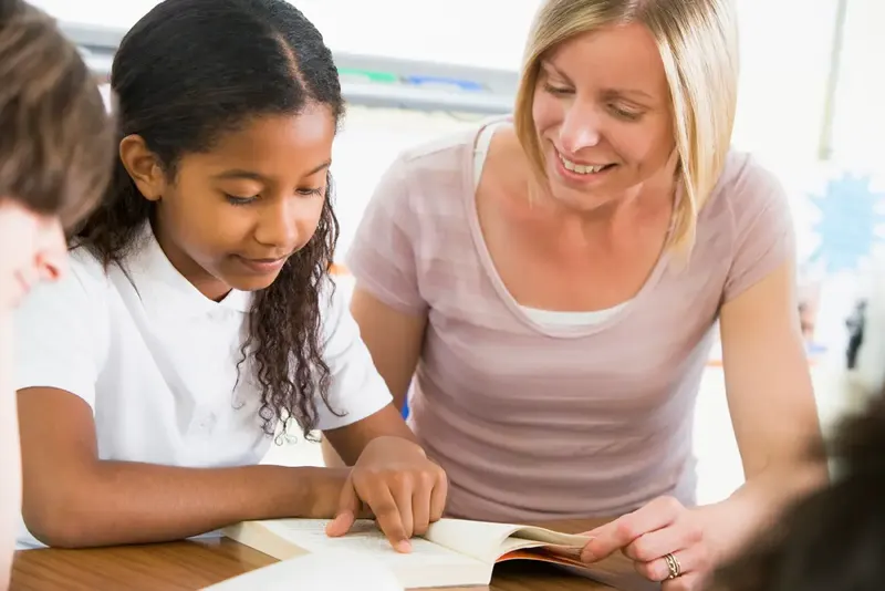 Eine Lehrerin beugt sich zu einer Schülerin und liest mit ihr in einem Buch